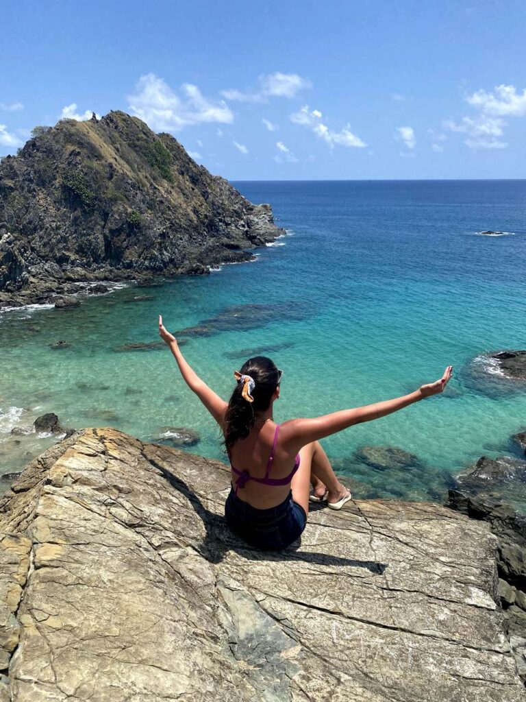 Lívia Pereira contempla de braços abertos sentada numa pedra num dia ensolarado o mar da Praia de Conceição em Fernando de Noronha