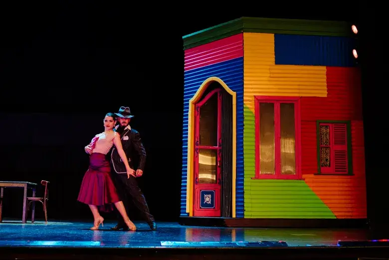 Casal dançando tango em Buenos Aires, num show em espaço fechado, o espaço conta com um cenário, no lado direito há uma casa colorida com porta e janelas, do lado direito uma cadeira e mesa