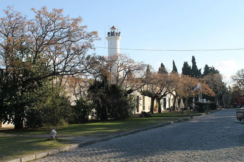 Centro histórico de Colônia do Sacramento no Uruguai num dia de sol, imagem de uma rua de paralelepípedos com muitas árvores e uma torre mirante ao fundo. Foto - Reprodução tripadvisor.com.