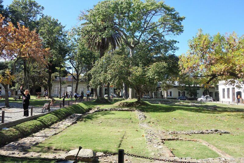 Plaza de Armas em Colônia do Sacramento no Uruguai. Imagem de um dia de sol forte, a praça tem muito verde formado por árvores e grama, ao redor tem uma série de construções históricas. Foto - Reprodução tripadvisor.com.