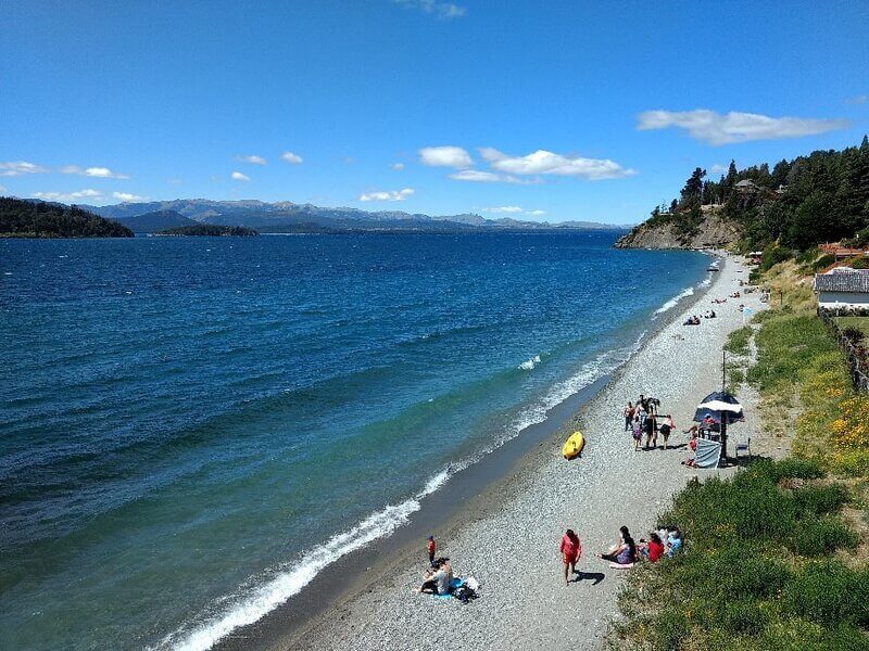 Orla da Playa Bonita em Bariloche na Argentina com alguns banhistas durante um dia de sol e céu aberto. Reprodução: Tripadivisor.