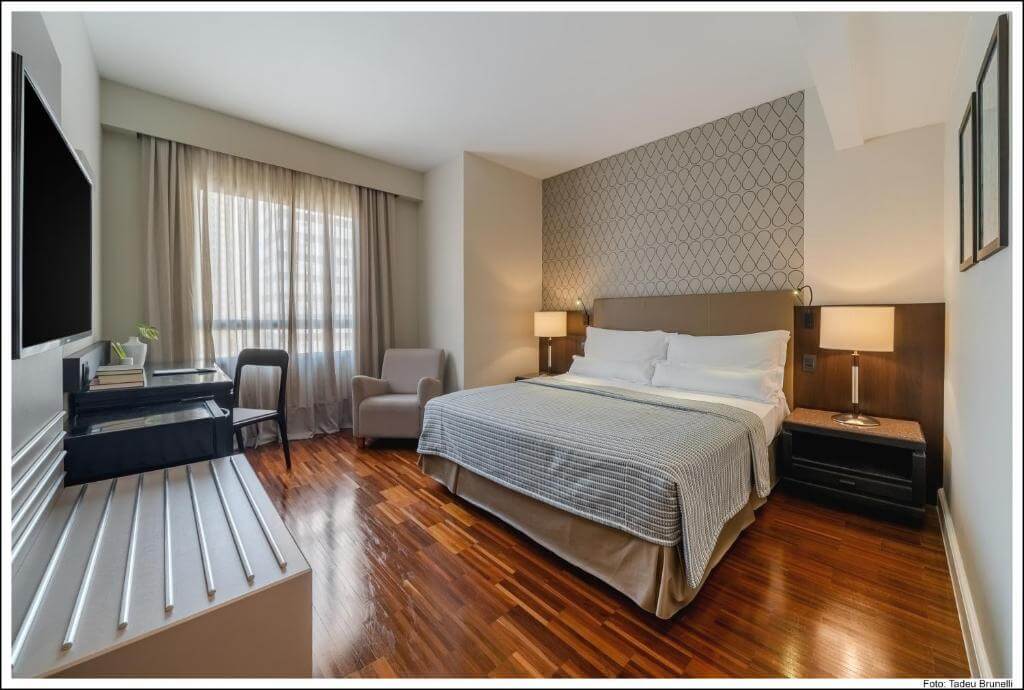 Quarto do Bourbon Curitiba Convention Hotel, em Curitiba no Paraná, com cama de casal, poltrona, mesa de escritório, televisão de tela plana, luminárias, mesa e escritório