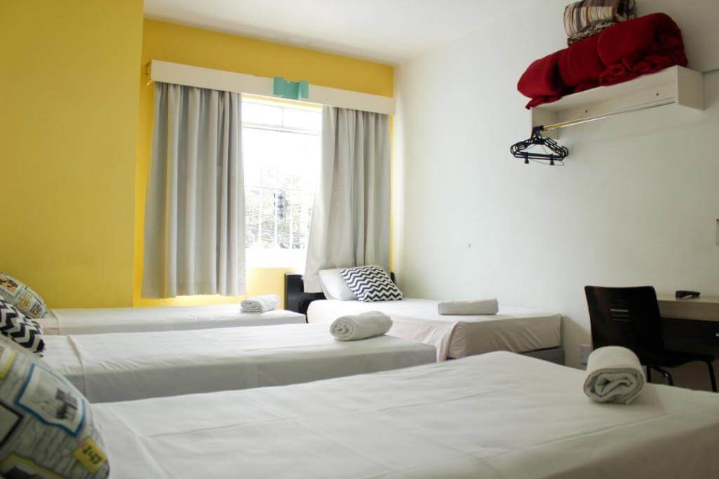 Quarto do Hotel Blumenau Centro, em Curitiba no Paraná, com camas de solteiro, armário, lençóis, mesa de escritório