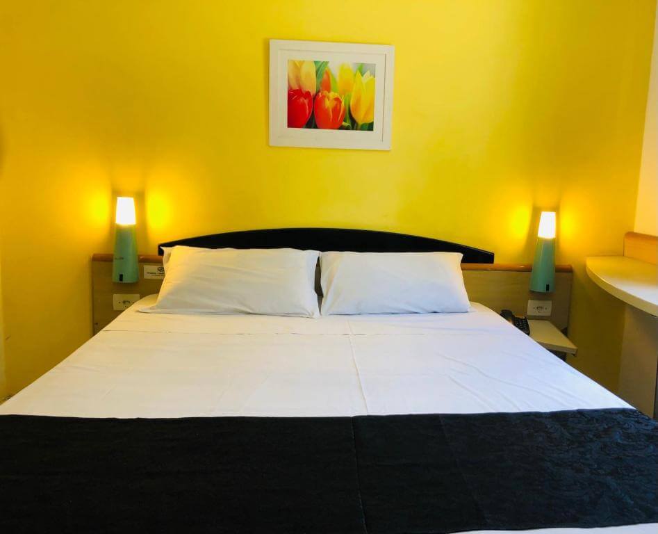 Quarto do Hotel Blumenau Centro, em Curitiba no Paraná, com cama de casal, travesseiro, luminárias e quadro