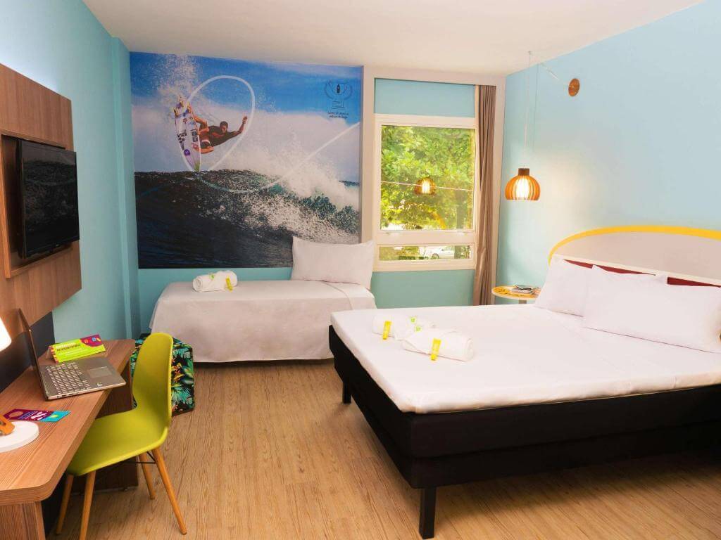 Quarto do Hotel Ibis Styles Enseada, em Guarujá no Litoral de São Paulo - SP, com cama de casal, mesa de escritório, tv de tela plana, puff e divã