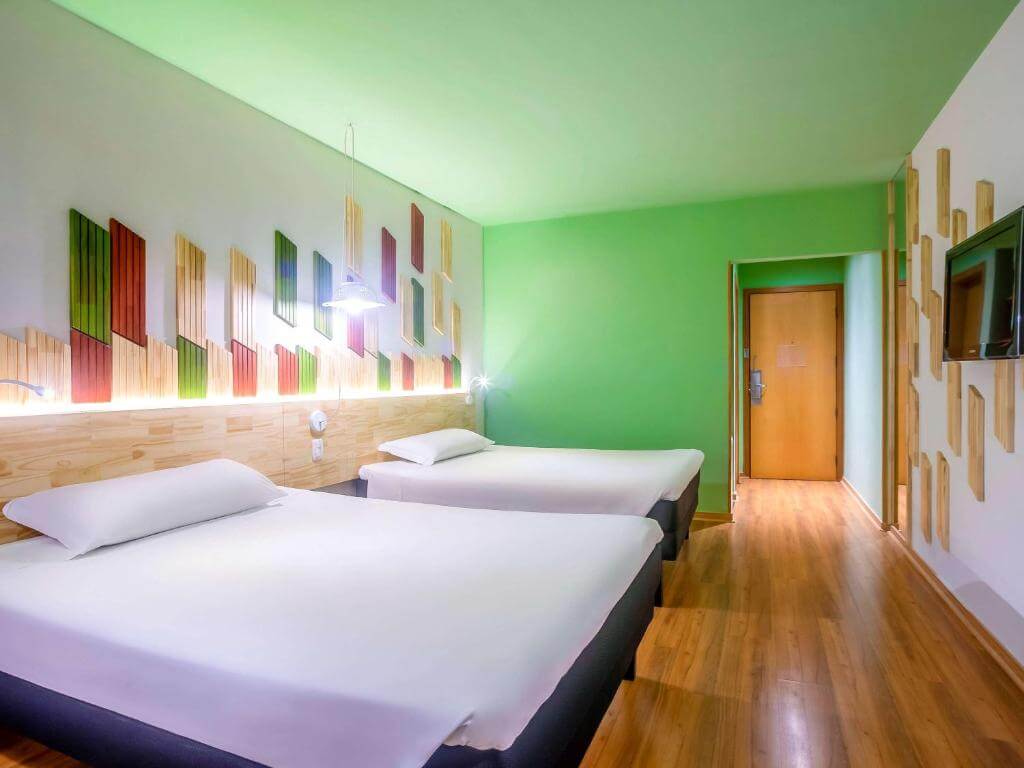 Quarto do Ibis Styles Curitiba Santa Felicidade, em Curitiba no Paraná, com duas camas de casal e uma televisão de tela plana