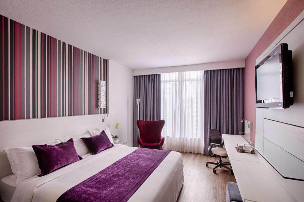 Quarto do Radisson Hotel Curitiba, em Curitiba no Paraná, com cama de casal, travesseiros e lençol na cor roxa, televisão de tela plana, poltrona na cor vinho, mesa de escritório e cadeira de escritório