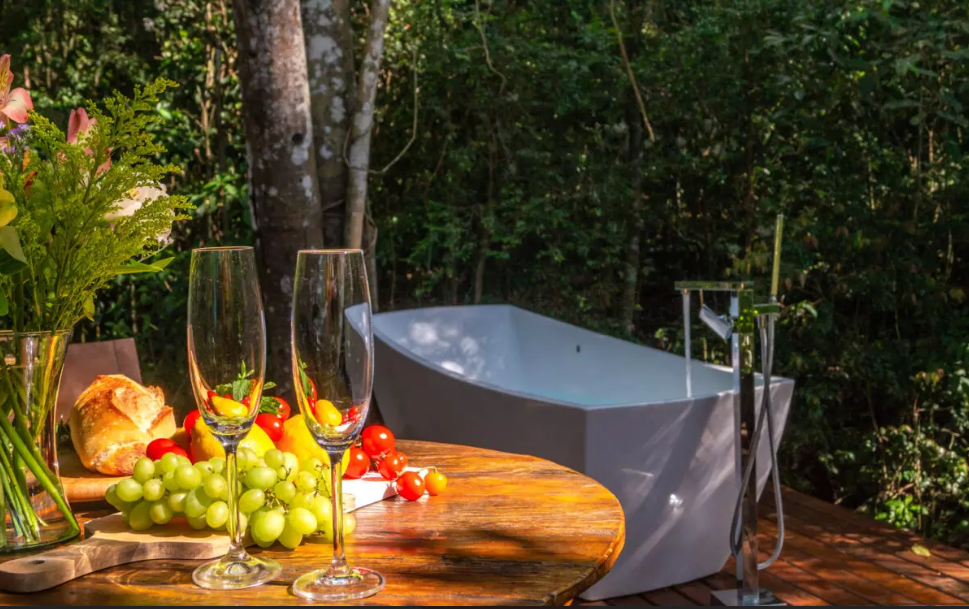 Banheira externa, com vista para a mata em Chalés românticos em São Roque. No detalhe, mesa com frutas e duas taças.