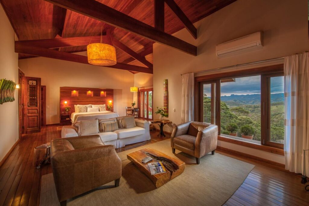 Quarto aconchegante, com luz baixa, detalhes em madeira. Cama de casal se compõe com uma sala de estar e vista das montanhas. 