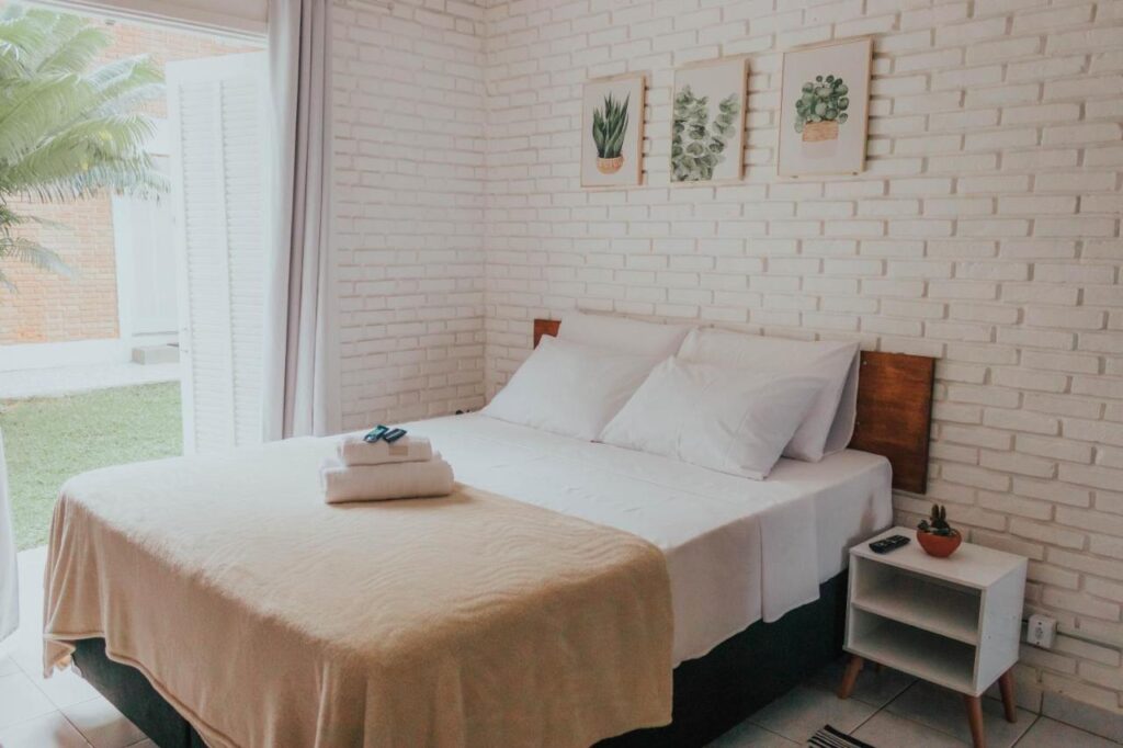 Quarto de casal com janelão para a área externa. Decoração clean em branco e detalhes marrons