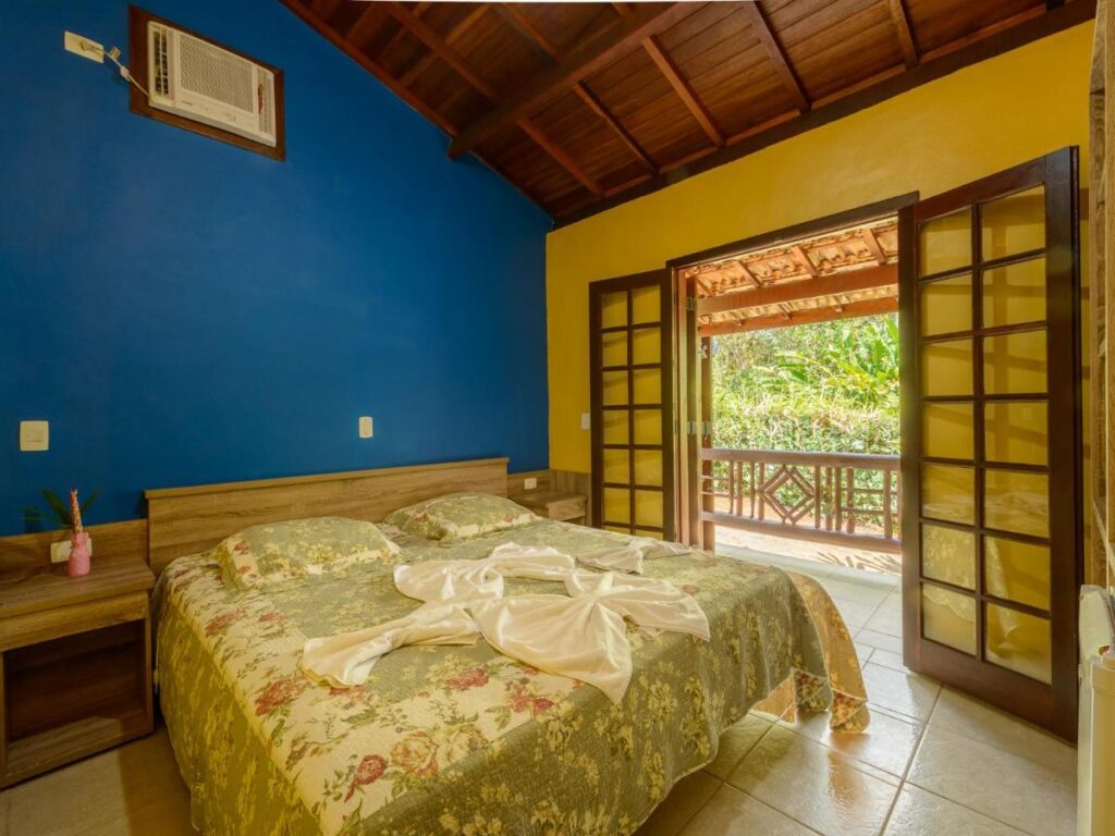 Quarto cama de casal, com parede em azul e janela porta balcão ao lado da cama, com vista para natureza