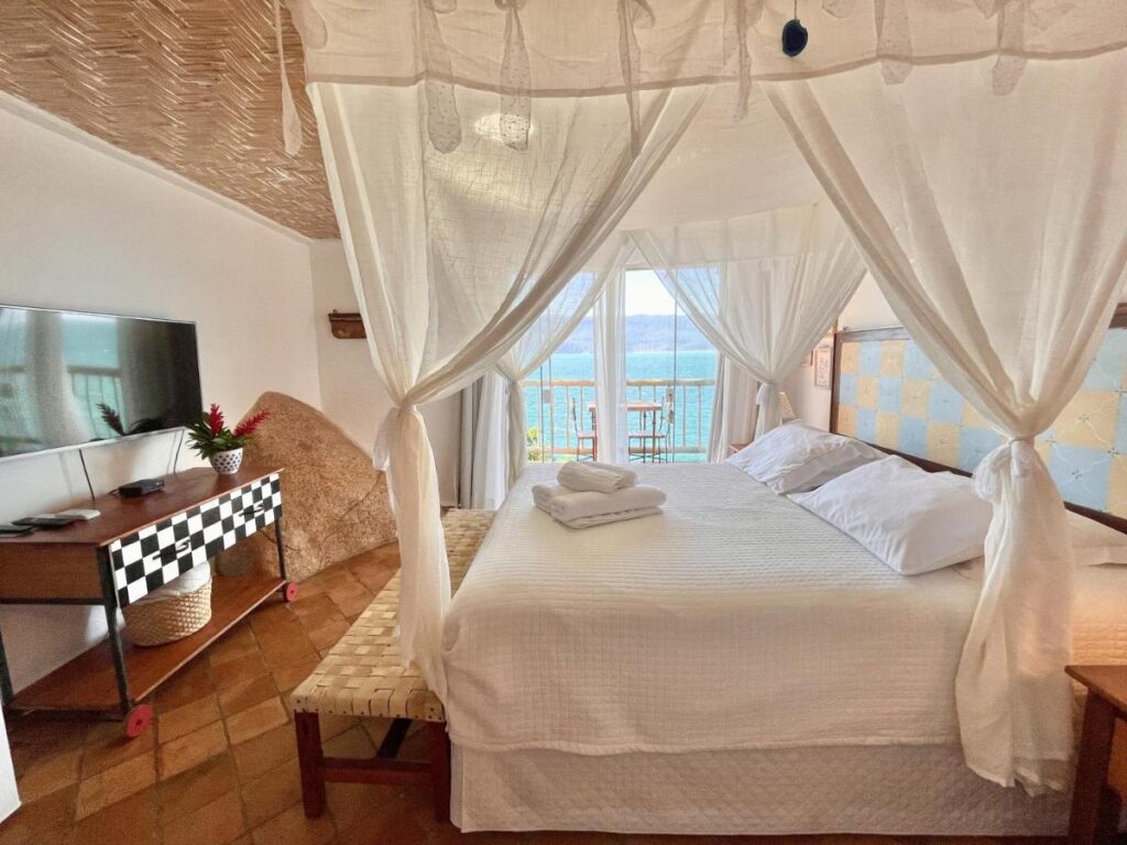 Quarto com cama de casal e mosquiteiro, ao fundo, o visual do mar pela varanda em hotéis de luxo Ilhabela